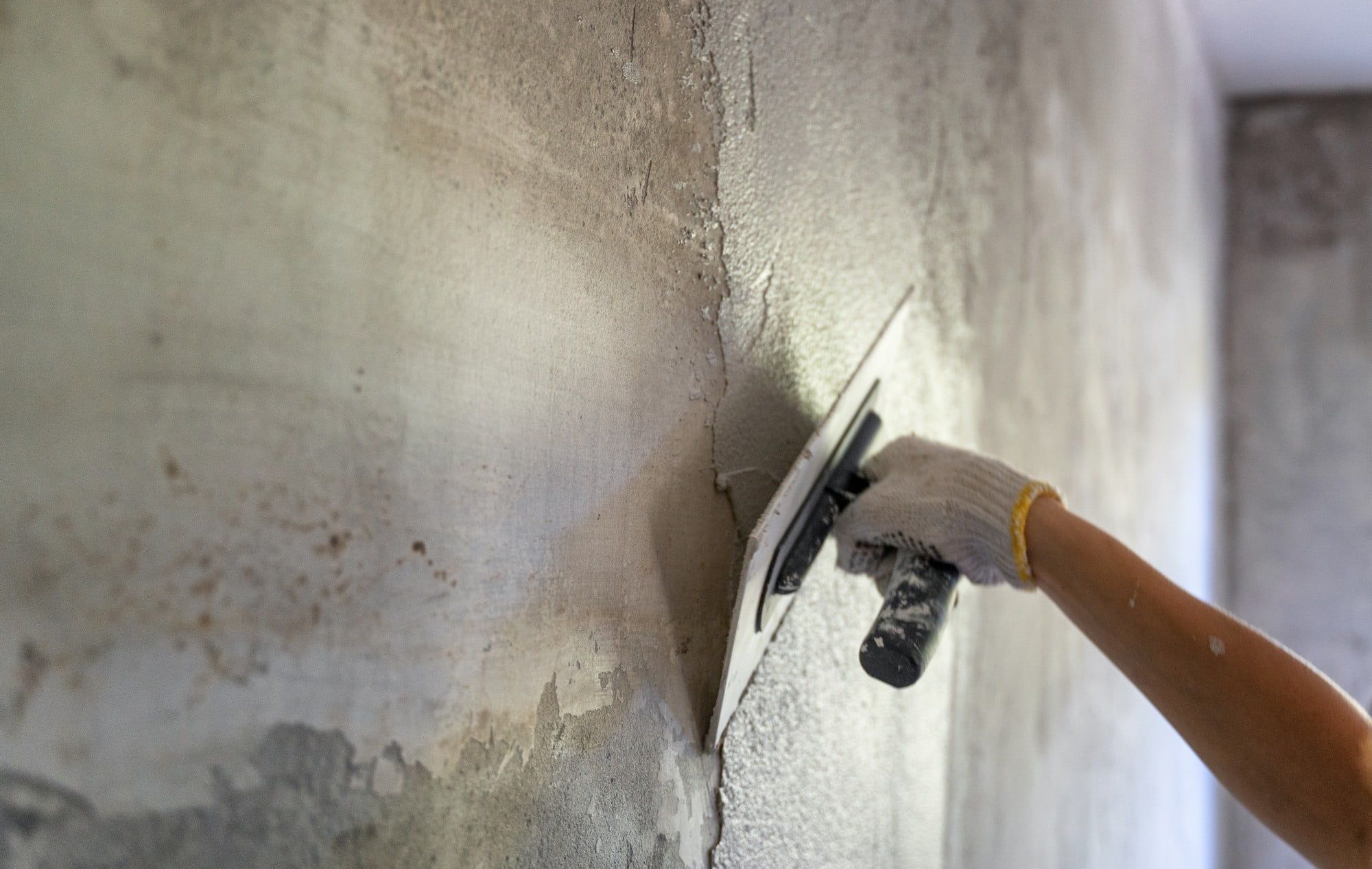 The image depicts a construction or renovation scene where a person's hand is applying plaster or spackle to a wall using a trowel. The person is wearing a glove, which suggests the work may require protection from rough materials. The wall appears to be unfinished or in the process of being repaired, with patches of rough and smooth areas indicating places where the plaster has been applied. The action captured is likely a step in the process of smoothing out the wall's surface in preparation for further finishing, such as painting or wallpapering.