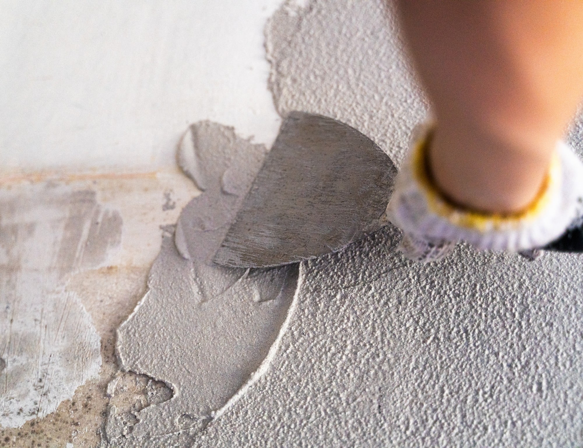 The image depicts a close-up shot of a hand wearing a glove, pressed against a wall with a texture that resembles raw plaster. The glove is made of a material that appears to combine elements of latex and fabric, offering both protection and flexibility. The plastered wall has a rough, unfinished look, with visible lines and irregularities characteristic of raw construction work. The lighting in the image is soft, casting gentle shadows that highlight the textures on both the glove and the plastered surface. The composition captures a realistic and tactile sense of touch, evoking themes of craftsmanship and manual labour.