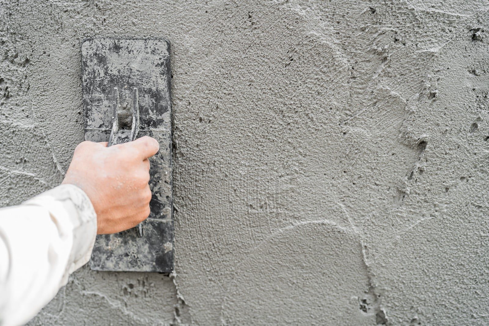 The image shows a person's hand holding a plastering trowel against a wall that is being plastered. The wall has a fresh layer of wet plaster, and it appears that the person is in the process of smoothing or applying the plaster to the wall. This is part of the process of finishing a wall, typically before it’s painted or otherwise finished. The trowel is coated with the plaster material, indicating it's been used to apply or spread the mix on the surface.