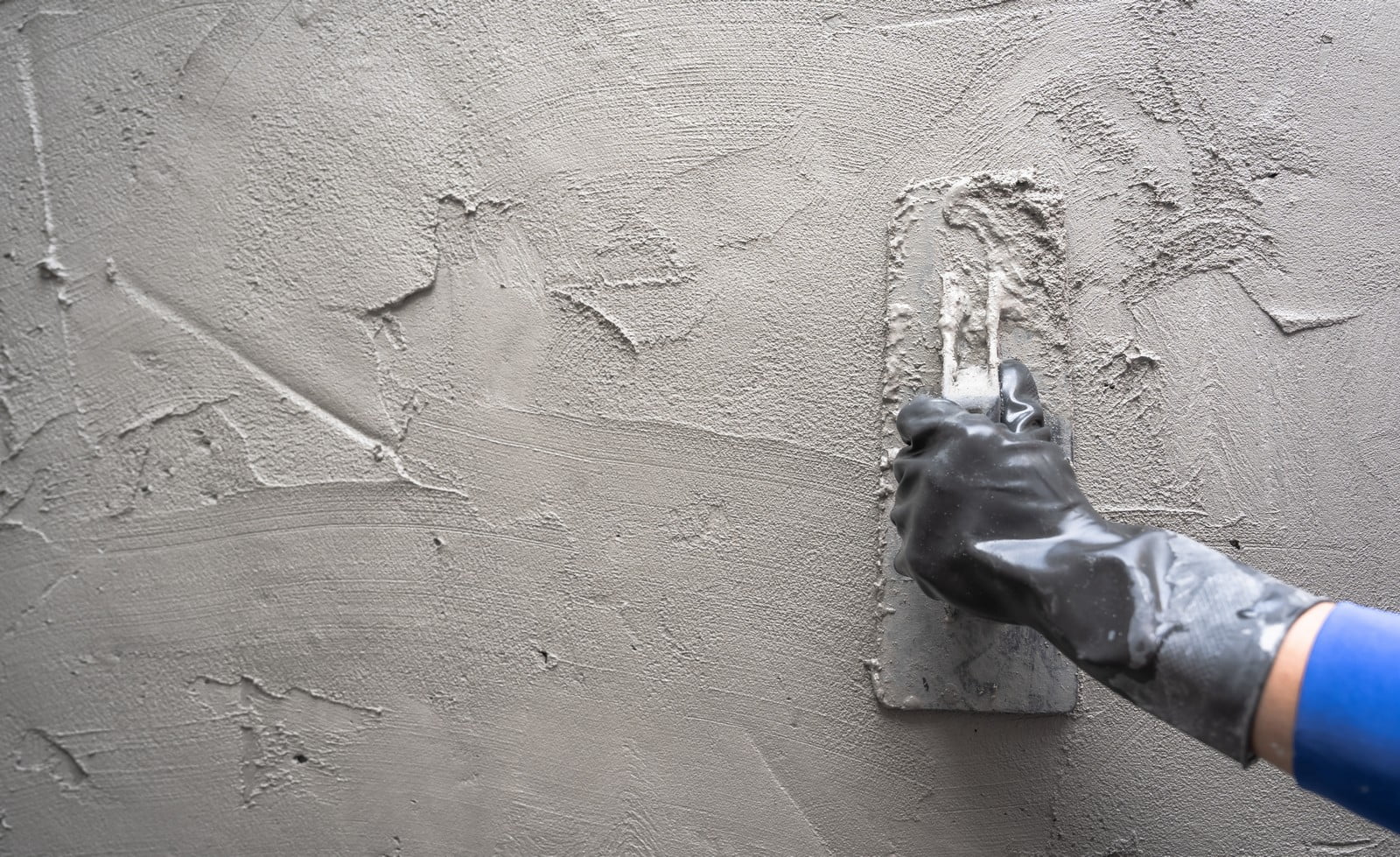 Sure, here is the detailed alt text description for the image:"A close-up photograph of a skilled hand plasterer applying smooth, wet cement plaster to a gray wall surface. The plasterer's hand, covered in a light layer of cement and wearing a protective glove, is shown holding a flat trowel at a slight angle, skillfully spreading the plaster in even strokes. The fresh layer of plaster appears seamlessly unified, creating a sleek, uniform texture across the wall. Sunlight filters into the scene from an unseen window, casting soft illumination and subtle shadows that highlight the smoothness of the newly applied plaster. The image captures the detailed craftsmanship and precision required in plastering work."