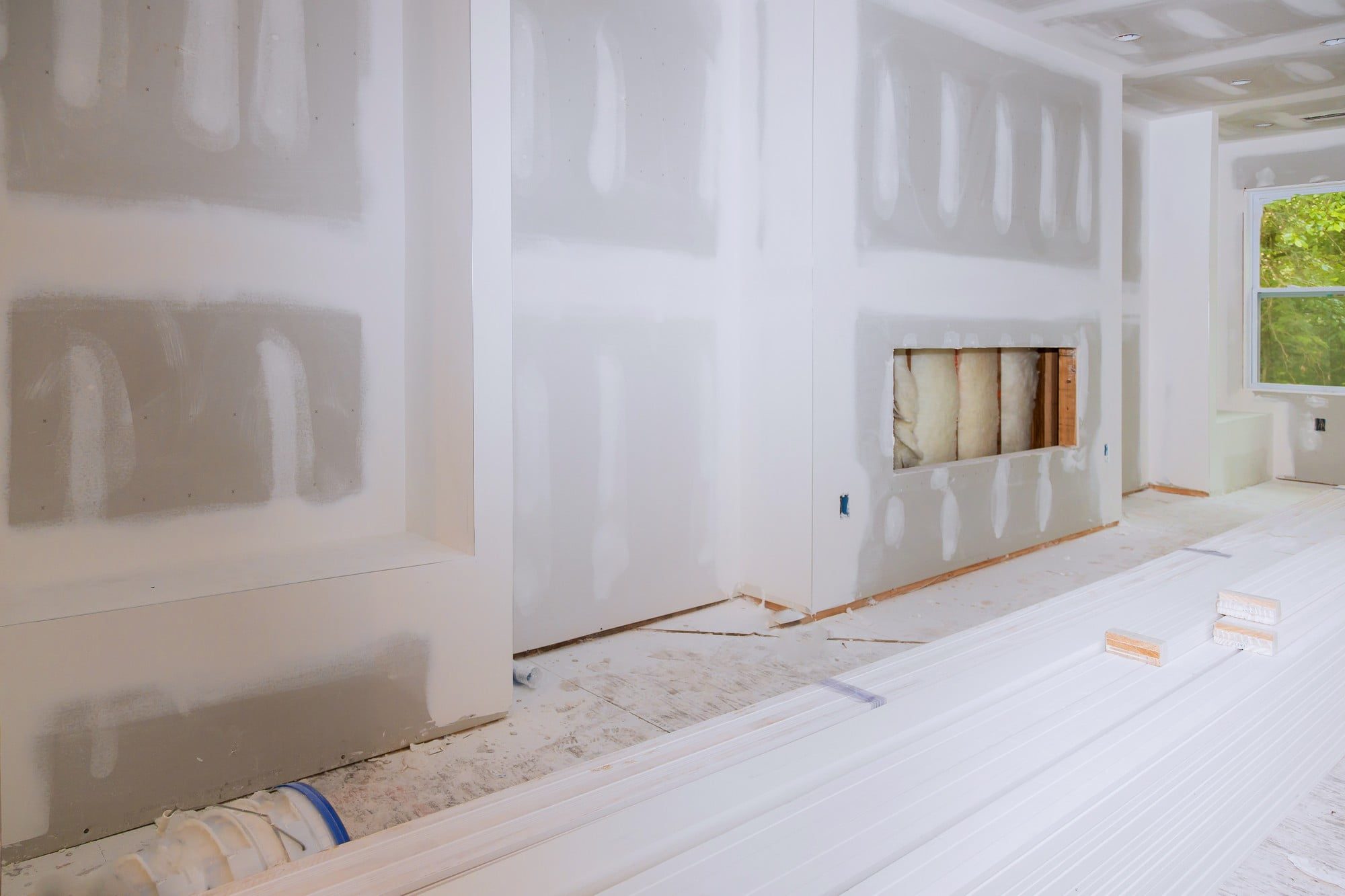 This image shows the interior of a room under renovation. The drywall has been installed on the walls, and joint compound has been applied over the seams and screw holes, which appears to be in the process of drying. Parts of the wall have been sanded, as evident from the smooth texture contrasting with the rougher areas covered in joint compound. There's an unfinished fireplace or some type of niche in the wall that is currently exposed, showing insulation material in the cavity.In the foreground, there are several lengths of white trim material or molding lying on the floor, likely planned to be installed along the baseboard or possibly around doorframes or windows. The floor is covered with protective material to prevent damage during the renovation process.The room seems to be well-lit, with natural light coming in from a window visible to the right. Electrical outlets are visible on the walls but not yet completed with cover plates. Overall, the image illustrates a typical stage in the home renovation process.