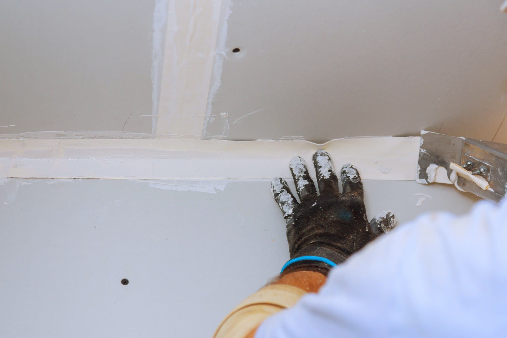 The image shows a close-up view of a person’s hand engaged in drywall construction or repair. The hand is wearing a black glove covered with white compound, indicating that this person is applying joint compound, also known as mud, to seams or gaps between sheets of drywall. To the right of the hand is a drywall taping knife, a tool used to spread and smooth the joint compound. The wall surface seems to be unfinished, with seams that have been taped and mudded, and screws slightly visible, which are typically used to affix the drywall to the framing behind it. The process depicted is typical for preparing a wall for painting or wallpapering after hanging drywall panels.