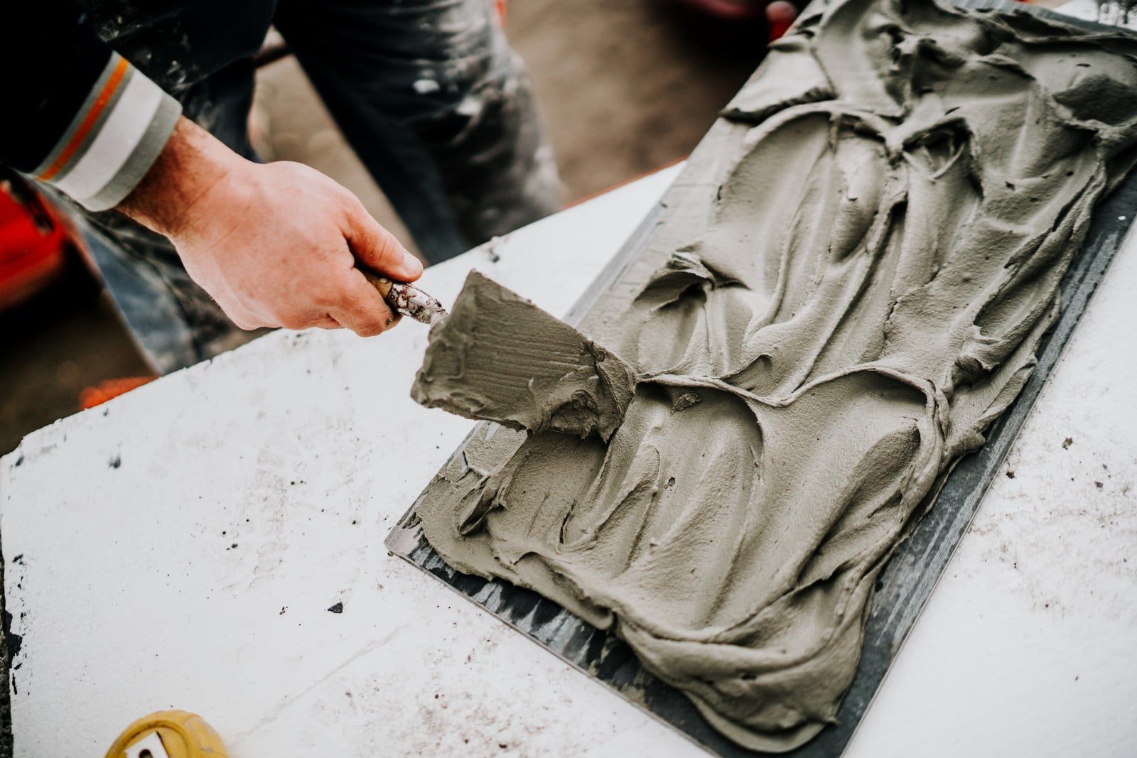 The image shows a person working with a trowel applying wet cement or plaster to a surface. It looks like they are either preparing a surface for tiling, plastering a wall or creating a decorative texture. The material being applied is thick and has been spread unevenly to cover the area, indicating that the work is in progress. The person's focus on the task at hand is evident from their grip on the tool and the methodical way the material is being spread.