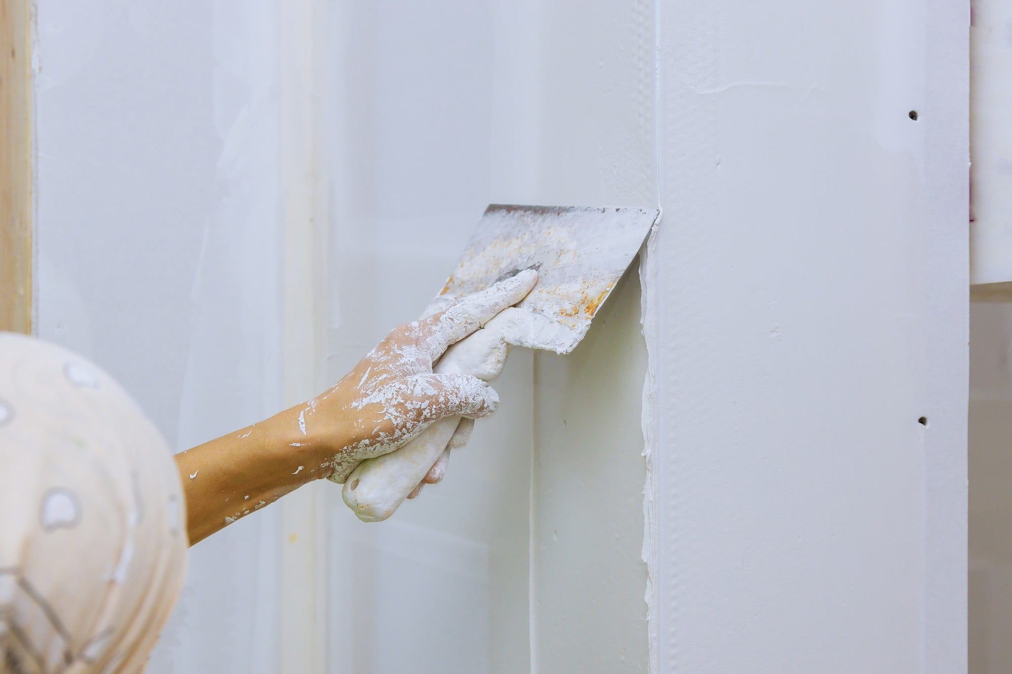 The image shows a close-up of someone's hand using a plastering trowel on a wall. The wall appears to have drywall installed with visible vertical metal or plastic beads or corners, which are typically used to protect the corners and create a clean line. The hand is covered in white dust, likely plaster or joint compound, indicating that the person is in the process of applying or smoothing the material on the wall, possibly as part of construction, renovation, or repair work. A safety helmet can be seen, suggesting adherence to safety protocols on the construction site.