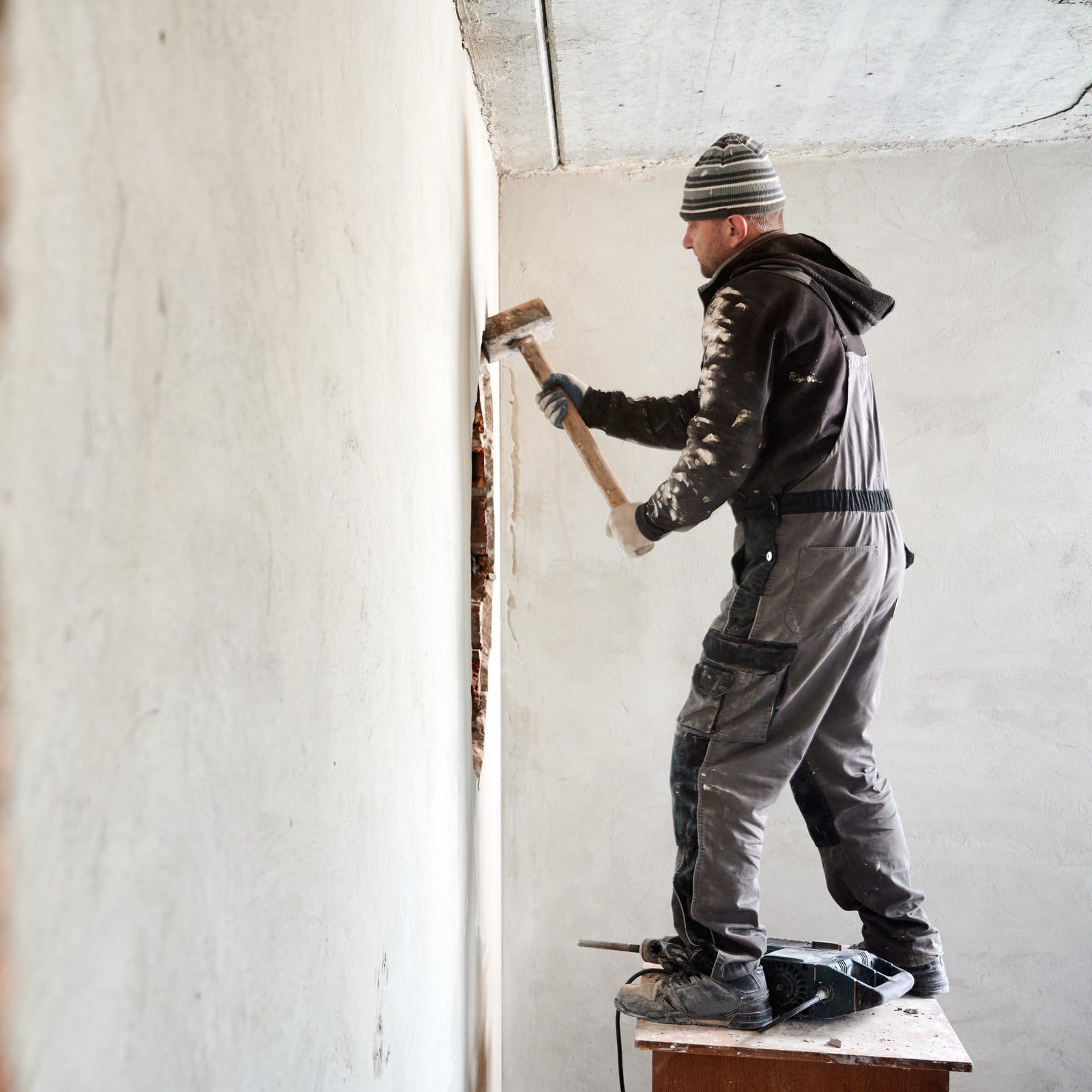 In the image, there is a person who appears to be engaged in some form of manual labour or renovation work. This individual is dressed in work clothes consisting of a jacket, pants, and a beanie hat, and is standing on what looks like a makeshift wooden platform or step. The person is wielding a sledgehammer and is in the process of swinging it towards a wall, likely in the action of demolishing or chipping away at the wall's plaster or structure. The environment suggests that this is an indoor setting, possibly within a building that is under construction or being renovated. The walls look partially stripped, indicating that the renovation process is ongoing.