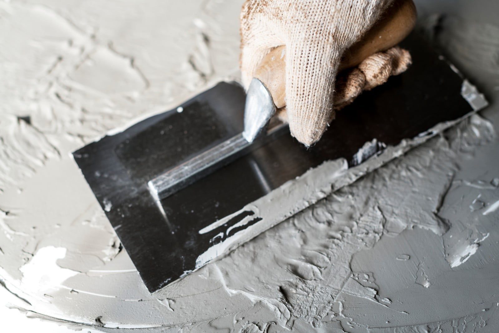 The image shows a close-up view of a person's gloved hand working with a plastering trowel. The trowel is being used to spread and smooth wet plaster or compound across a flat surface, which appears to be either a wall board or a practise board for plastering. The plaster is gray in colour, and it seems to be freshly applied, given its wet appearance. The surface beneath the plaster is dark, possibly made of a plastic or metal material, which contrasts with the light gray of the plaster. The person is likely wearing a protective glove to keep their hand clean and to protect it from any potential irritants in the plaster.