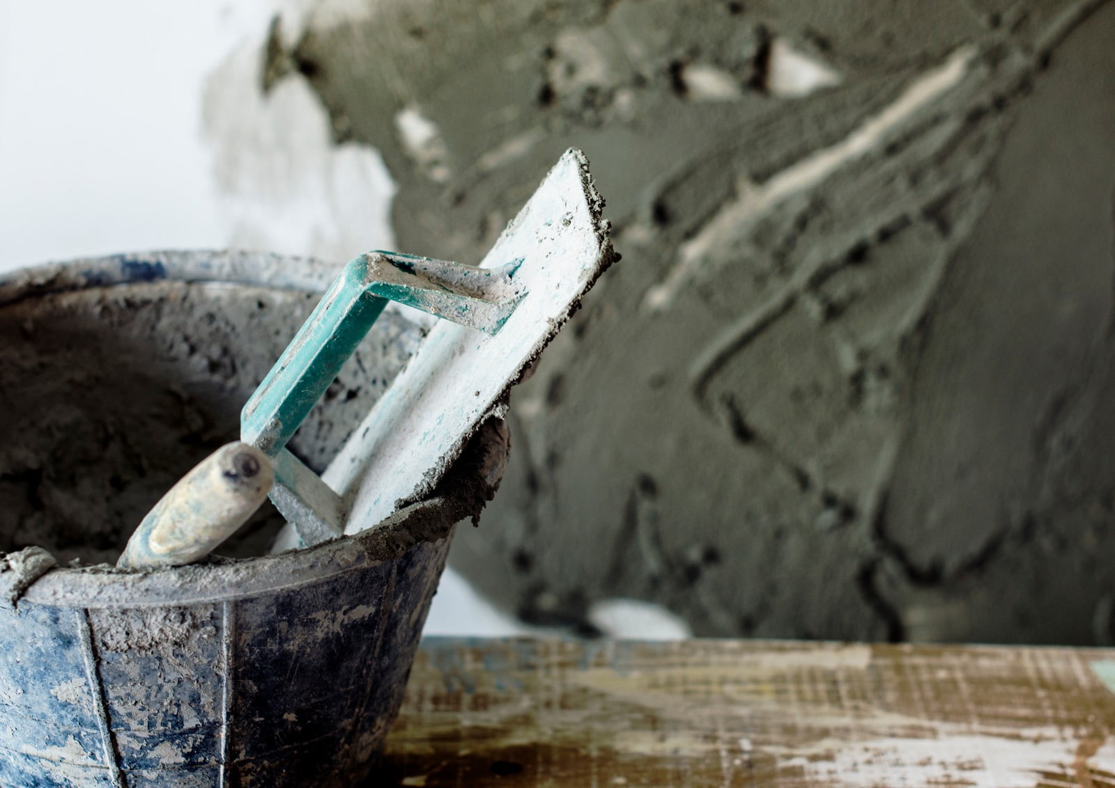 The image shows a close-up view of construction tools and materials. There is a blue bucket that appears to contain wet cement or plaster, with a plastering trowel leaning against it. The trowel has a greenish handle and is coated with the same material as in the bucket, indicating it has been used. Behind the bucket, there is a wall with a freshly applied coat of the same substance, showing trowel marks where the plaster has been spread. The foreground shows a wooden surface that's also speckled with the plaster, perhaps a work table or support where the bucket has been placed. The image captures the process of wall plastering during a construction or renovation project.