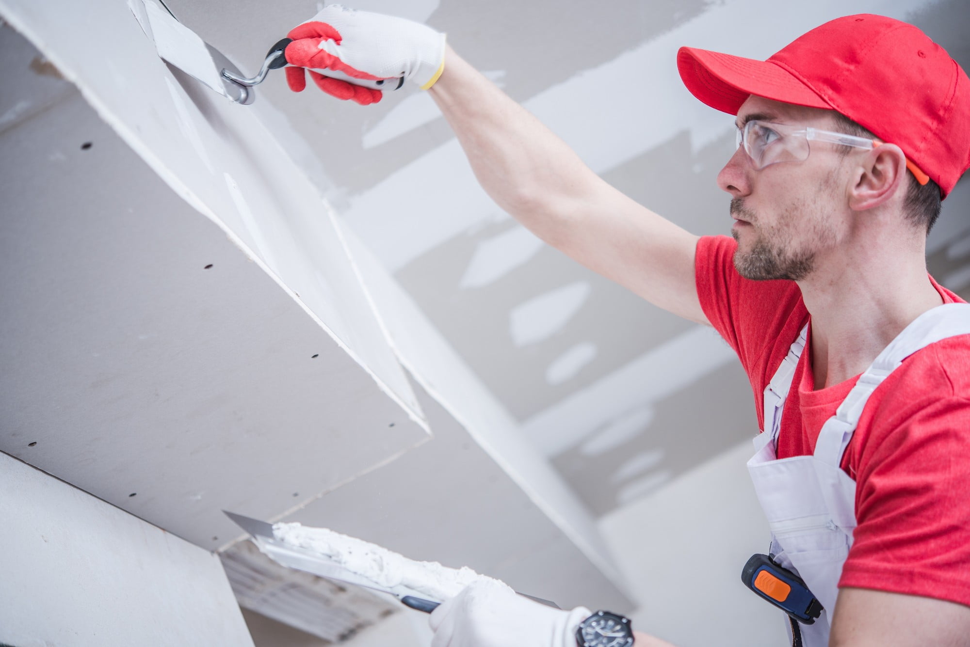 This image shows a person applying joint compound or spackle to a seam in drywall. The person is dressed in work attire that includes a red cap, safety glasses, and a white and red shirt. They are wearing gloves and a tool belt, indicating that they are a professional or skilled worker in the process of finishing walls, likely in preparation for painting or wallpapering. The environment suggests that it's an indoor setting, possibly under construction or renovation.