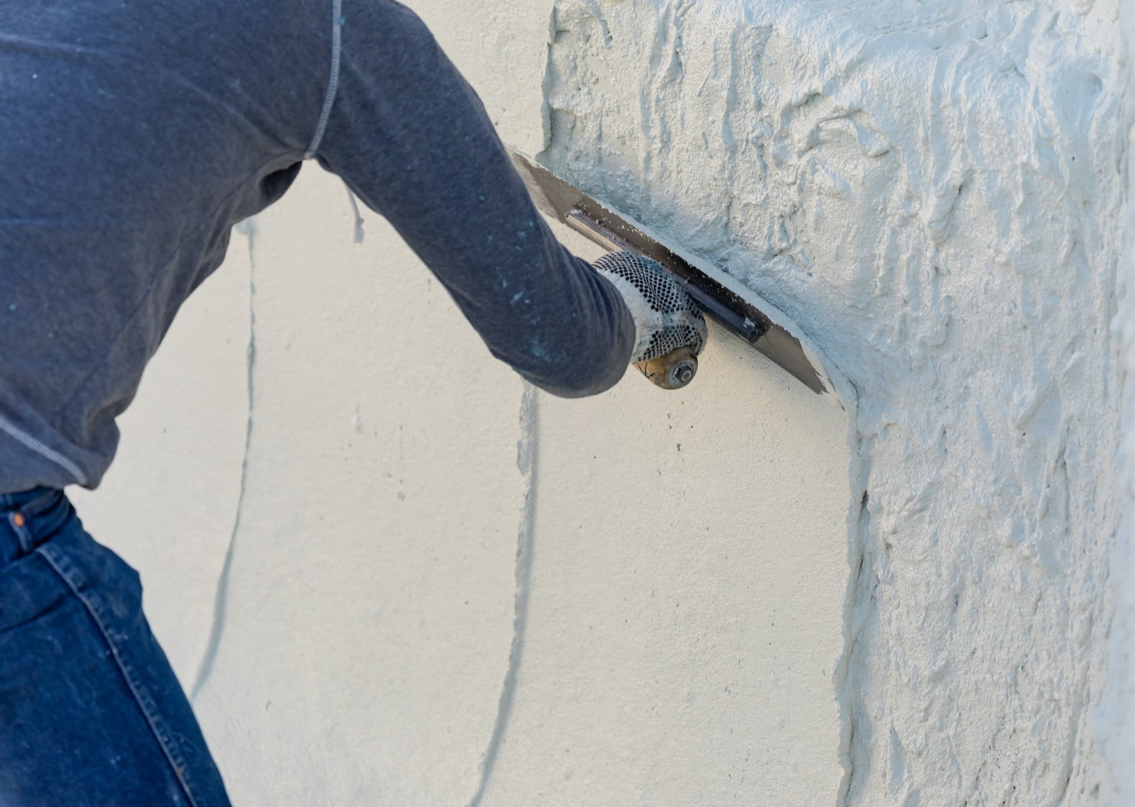 The image shows a person in the process of applying or smoothing what appears to be plaster or stucco to a wall with a trowel. The wall has a textured surface, and the person is wearing a gray long-sleeve shirt with jeans, and they have protective gloves on their hands. The work seems to be at an outdoor location, given the brightness and shadows indicating sunlight. The focus is on the action of applying the material to the wall, highlighting a moment of manual labour and construction work.