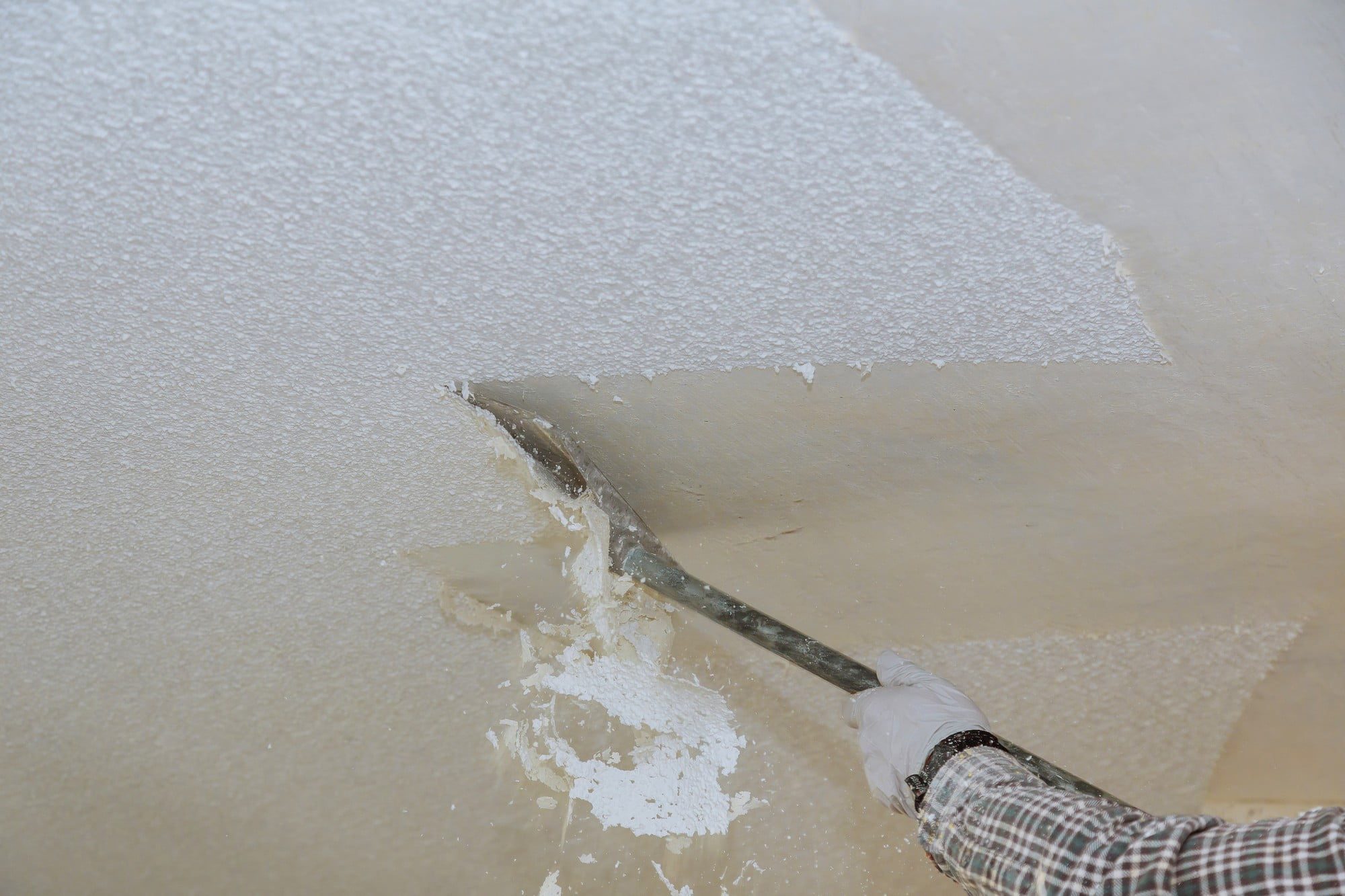 The image shows a person using a scraping tool to remove a layer from a floor. The floor appears to be partially covered with a white, textured surface that is being scraped away to reveal the underlying material. The person is wearing a glove for protection, indicating that this may be a construction or renovation task. It looks like they are in the process of removing old floor covering, such as adhesive residue or a material like paint or epoxy. The scraping tool seems to be a flat-edged tool specifically designed for such jobs.