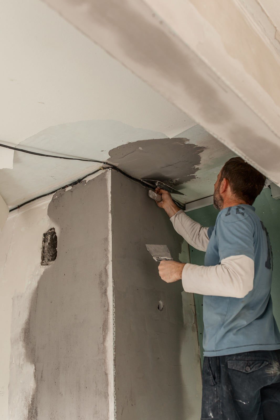 The image shows a person applying plaster on a wall. The individual appears to be in the process of smoothing out the plaster to create a flat surface and possibly preparing the wall for painting or wallpapering. They are using a hand tool, which looks like a plastering trowel or spatula, to evenly distribute and smooth the plaster. The person is dressed in casual work attire and seems to be focused on the task at hand. The wall and ceiling look incomplete, likely in a room under construction or renovation.