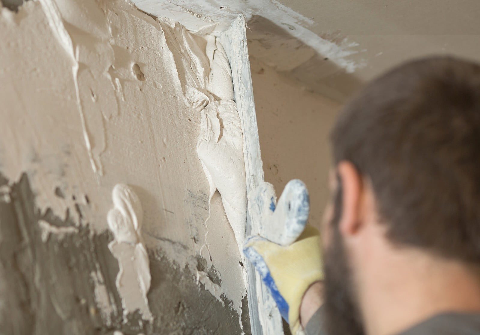 The image shows a person applying plaster or joint compound to a wall or ceiling corner with a tool, presumably a trowel or spatula. The individual is wearing protective gloves and is in the process of smoothing the material to create a seamless surface. You can see excess plaster squeezed out as the tool presses into the corner, indicating the person is likely doing finishing work related to construction or renovation, such as preparing the area for painting or wallpapering. The focus of the image is on the work itself, highlighting the texture of the plaster and the method of application.