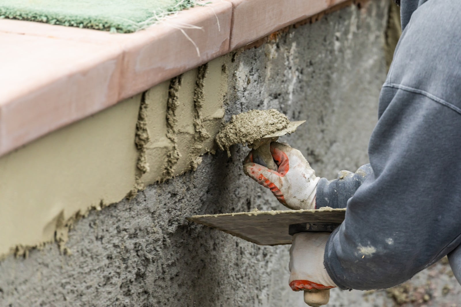 The image depicts a construction scenario where a person is applying wet cement or mortar to a vertical surface, which appears to be part of a wall under construction or repair. The individual is using a trowel to spread the material and has a hawk (a flat tool used to hold plaster or mortar) in their other hand. They are wearing protective gloves and a long sleeve shirt, indicating adherence to safety measures while working. The focus is on the worker's hands and the immediate task. You can see part of the building substrate, which has been coated with an underlayer before the current layer of mortar is being applied.