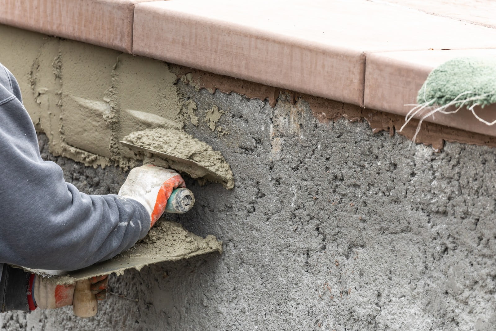 This image shows a construction worker applying wet cement or plaster to a wall using a trowel. The worker is wearing gloves and is in the process of smoothing the material onto the vertical surface. The cement is being applied under what appears to be a ledge or sill, possibly as part of the process of installing or finishing the ledge. The specific techniques and materials used will depend on the requirements of the building and the finishes desired. The image captures a common process in construction where masonry or plaster is used to create smooth or textured surfaces on walls.