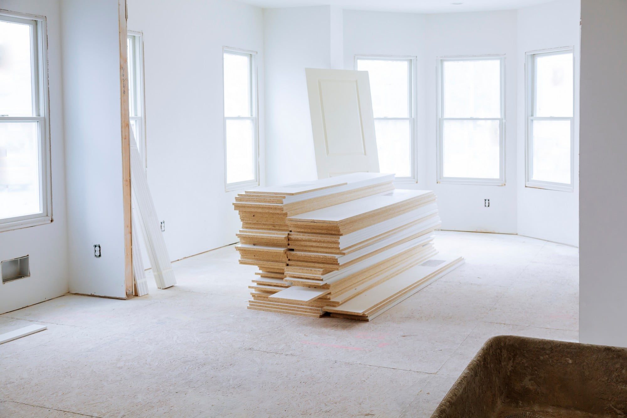 The image shows an interior space that appears to be under construction or renovation. There are several features visible:1. The floor is covered with subflooring material, possibly plywood or OSB (oriented strand board), which suggests that final flooring has not yet been installed.
2. The walls are painted white and look to be finished.
3. There are multiple white-framed windows providing natural light to the space.
4. A stack of white door panels or perhaps precut wall panels is in the centre of the room, indicating ongoing installation work.
5. Electrical outlets are visible on the walls, but without cover plates, which is common in spaces that are not yet fully finished.
6. There is a pile of narrow wooden planks or trim leaning against the left wall.
7. There are faint markings on the subfloor, possibly for laying out walls or fixtures.
8. In the bottom right corner, there's a bit of what looks like a wheelbarrow or a construction cart, likely used to transport materials.The overall impression is of a light, airy space midway through the renovation process.