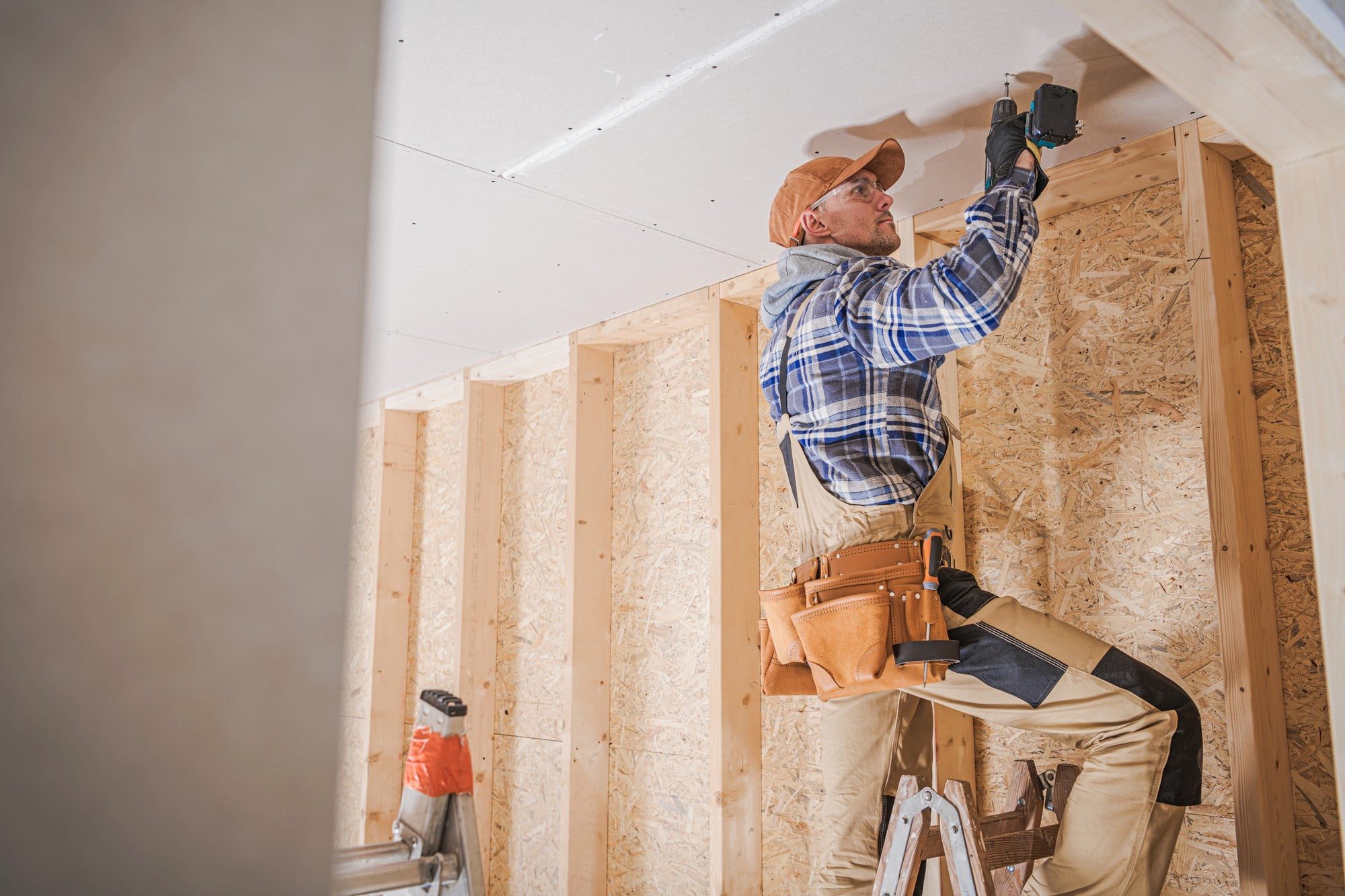 In the image, there's a person working on a construction site or in a building undergoing renovation. The individual is wearing a plaid shirt, a cap, and has a tool belt around the waist, which holds various tools and supplies. The person is using a powered screwdriver or drill to affix a sheet of drywall to the ceiling structure. Below the person, there is a stepladder, indicative of the fact that the worker used it to reach the ceiling. The walls behind the person are constructed from wooden studs and covered with oriented strand board (OSB), which is often used to sheath walls in residential or light commercial construction before the final wall finish is applied. The environment looks like an interior space that is in the midst of being finished or remodeled.