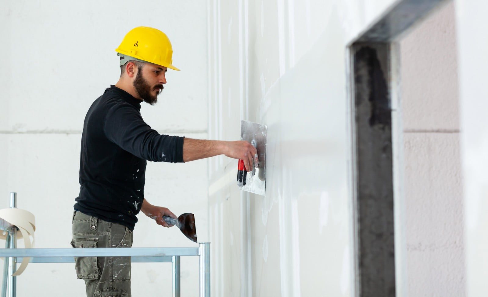 This image shows a construction worker or tradesperson applying plaster or joint compound to a wall. He is using a trowel and looks focused on smoothing the material over the surface. He's wearing a yellow hard hat, which is standard safety equipment on construction sites, and his clothes have splatters of paint or plaster, indicating active work. The environment appears to be an indoor construction or renovation site, given the unfinished surfaces and the scaffolding, which suggests he's working at height or needing to reach a higher part of the wall.