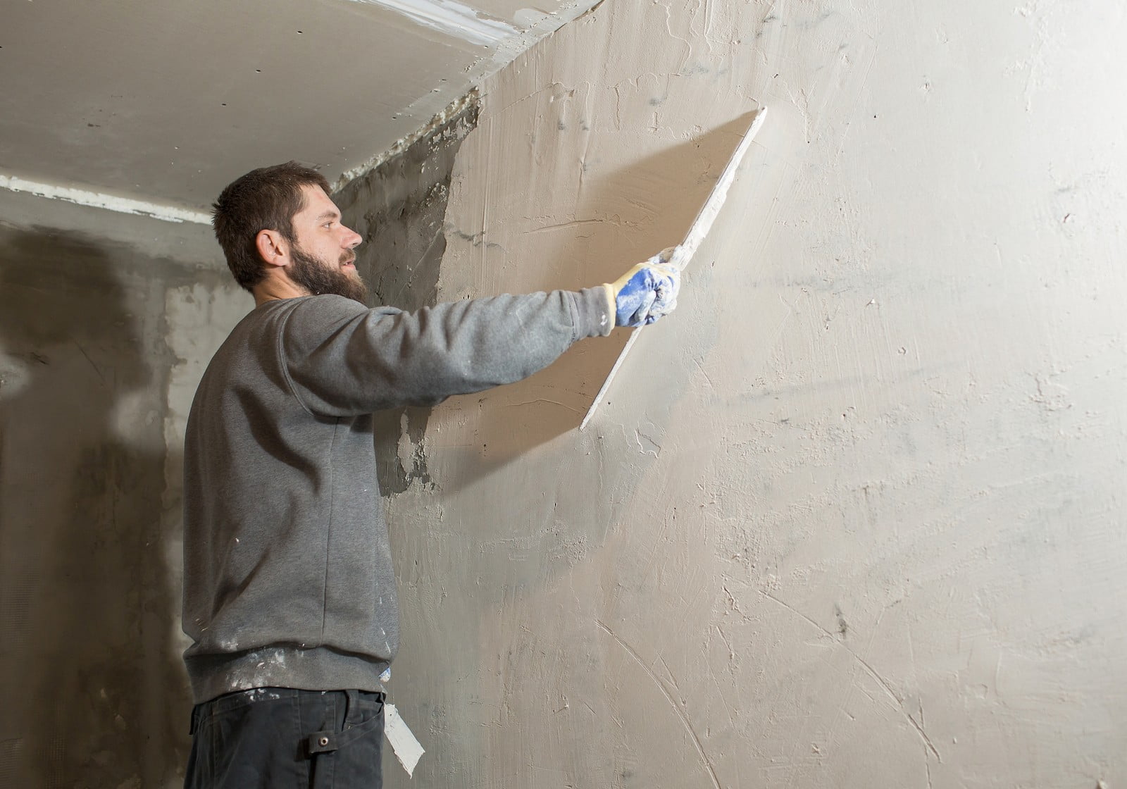 The image shows a person applying plaster to a wall with a large trowel. The person appears to be in the process of smoothing the surface, which is common in construction or renovation to prepare the wall for painting or wallpapering. The wall looks partially covered with wet plaster, and the floor is protected with a drop cloth. The person is dressed in casual workwear with gloves, indicating that they are observing safety measures while doing the plastering work.