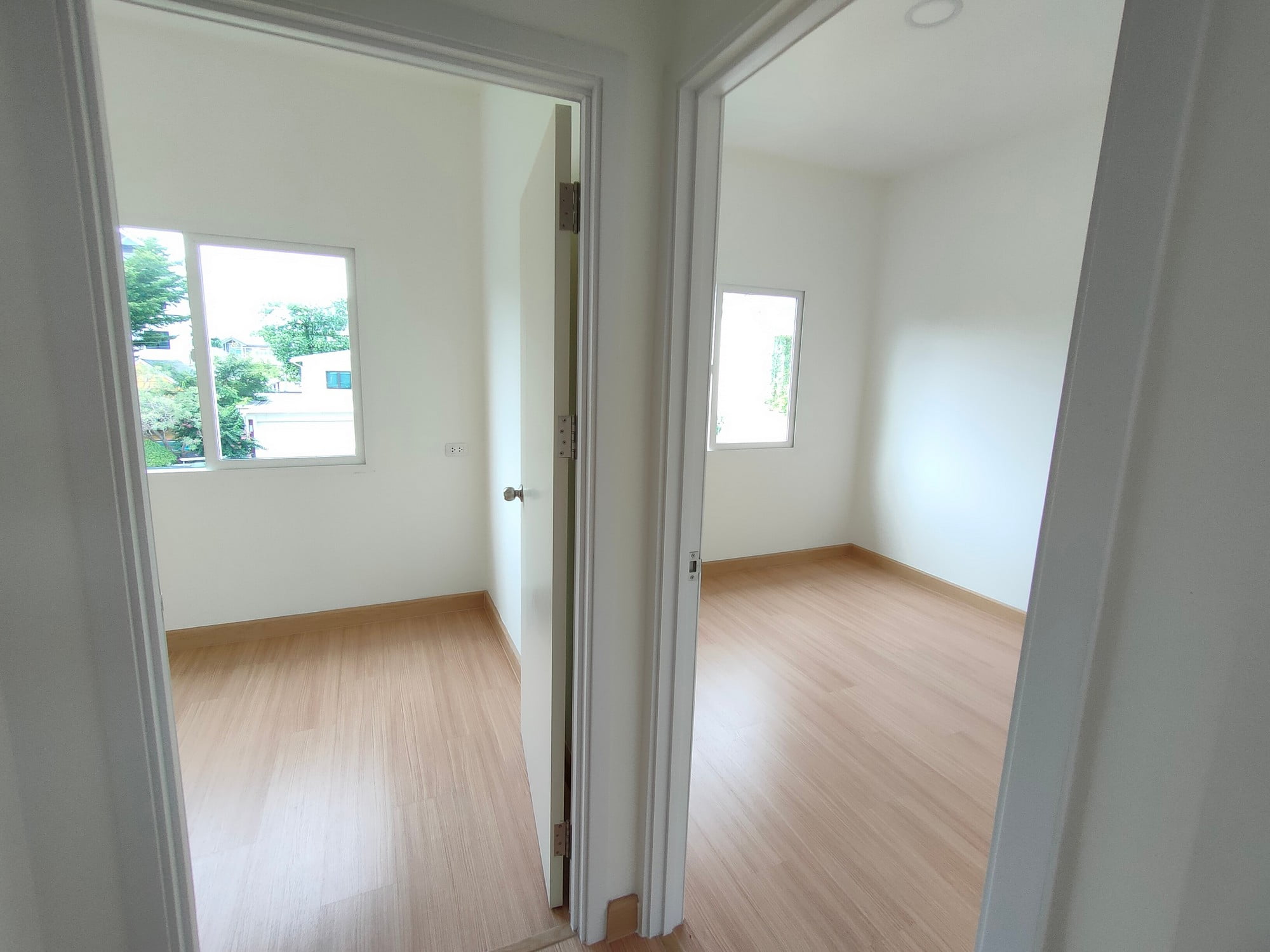 The image shows a bright, empty room with a wooden floor. The room has white walls and gets plenty of natural light from the two visible windows, which offer a view of the outside environment with greenery and other residential structures. There's a white open door providing entry into the room, with the door handle suggesting that this is an interior door. This looks like it could be a residential space, such as a bedroom, office, or living room in an flat or house that is either newly constructed or has been recently cleared out. The simplicity and cleanliness suggest it might be ready for furnishing or showing to potential tenants or buyers.