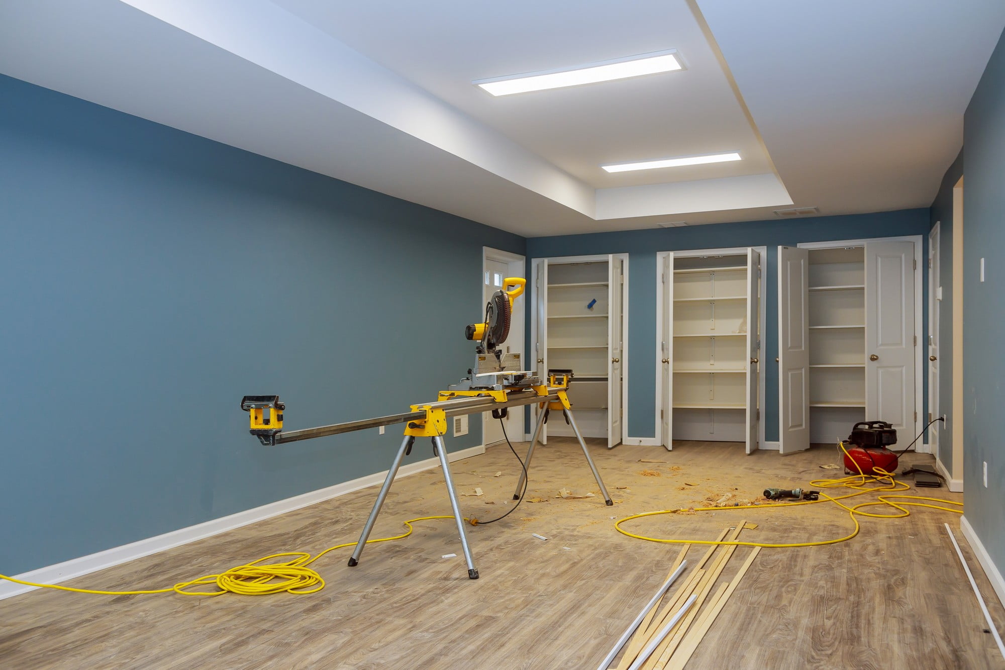 This image shows a room undergoing renovation or construction work. In the centre, there is a yellow compound miter saw placed on a stand, which is typically used for making precise crosscuts and angle cuts. There is also some sawdust on the floor, indicating recent use of the saw.On the floor, we see a coil of yellow extension cord and some wooden planks or trim pieces that might be used for the baseboards or framing. There's a vacuum cleaner to the right, possibly for cleaning up the construction debris. The room includes built-in white cabinets or closets on the right side, and the walls are painted in two shades with the bottom part a darker blue and the top part a lighter shade, separated by a white chair rail.The ceiling has modern rectangular LED light fixtures, providing bright illumination to the space. The floor has laminate or vinyl wood-look flooring. The general state of the room suggests active construction or remodeling, but no people are visible in the shot.