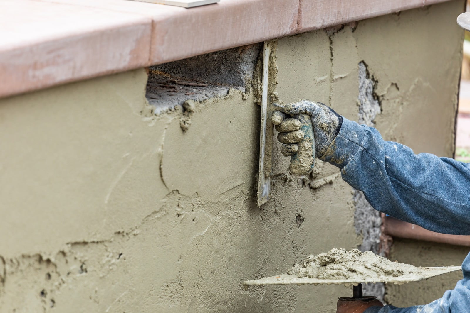 The image shows a person applying wet cement or stucco to a vertical surface, which appears to be an outdoor wall or structure. They are wearing a heavy-duty glove and are using a trowel to smooth the material onto the surface. Some of the underlying structure, possibly consisting of metal lath or mesh, which provides support and adhesion for the cement, is exposed. The person is likely engaged in construction, masonry, or repair work to finish or repair the wall surface. There are also some wooden elements visible at the top of the image, suggesting a framed area or a ledge.