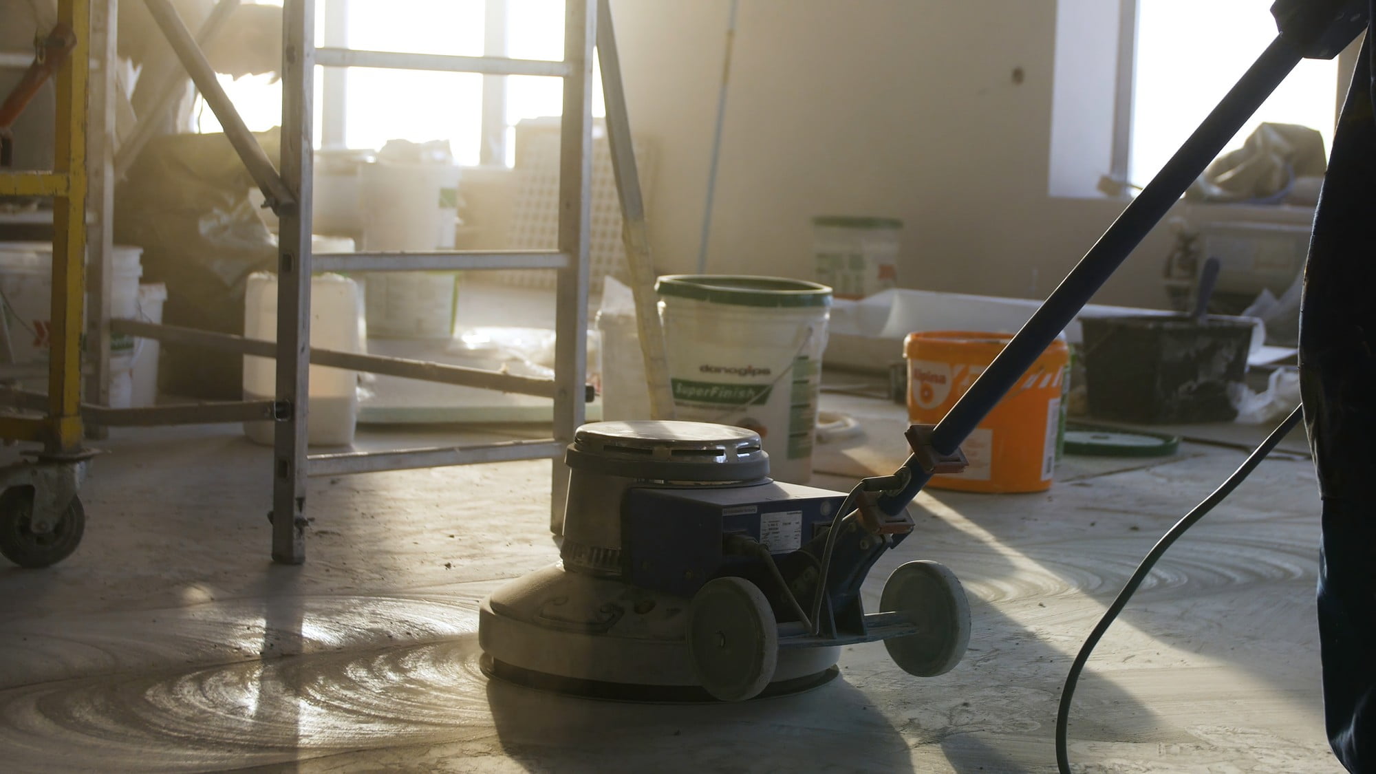 The image features a construction worker actively engaged in polishing a mortar floor at a construction site. The worker is wearing standard safety gear, including a white hard hat, a high-visibility yellow safety vest, and sturdy work trousers. He is kneeling on one knee as he operates a floor polishing machine, guiding it with diligent focus. The machine appears to be industrial-grade, with a large circular base and a sturdy handle, designed for efficiently smoothing and finishing mortar floors. The background reveals a partially constructed building, with exposed concrete walls and metal scaffolding indicating that the structure is still under development. The overall atmosphere is one of industrious activity, capturing a moment of precision and craftsmanship essential to the construction process. The lighting is natural, suggesting the photo was taken during the daytime.