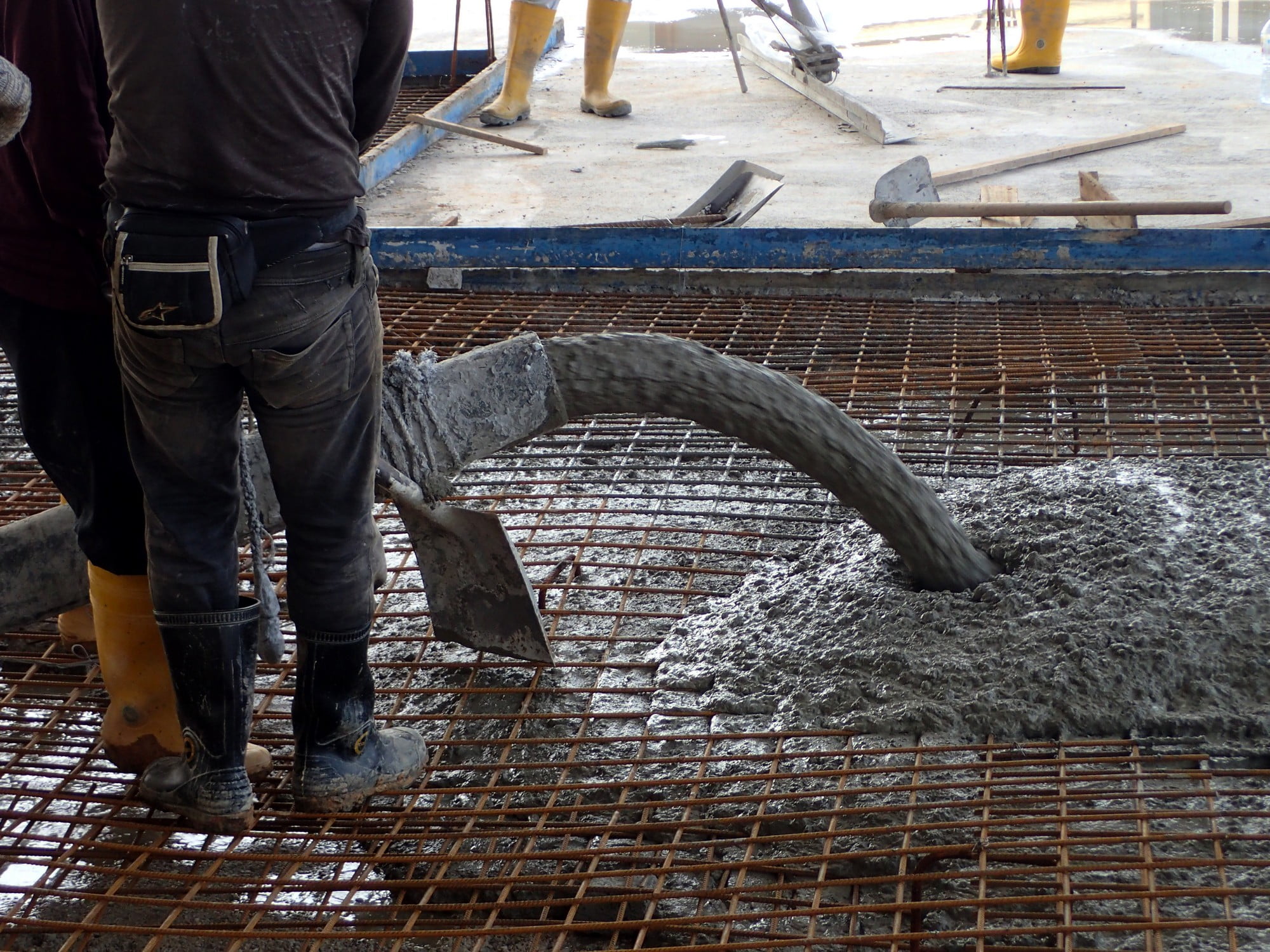 The image depicts a construction worker in the process of pouring concrete for a building foundation. The worker is wearing traditional safety gear, including a bright yellow hard hat and an orange reflective safety vest, which are standard for construction site safety. He is holding a large, thick concrete hose, which is actively dispensing wet concrete into a prepared rectangular form on the ground. The liquid concrete is grey and appears fresh and viscous. The backdrop features wooden and metal frameworks, signifying an in-progress construction zone. Various construction materials, including reinforcing steel bars and wooden planks, are scattered around the area, highlighting the industrial nature of the site. The day appears to be clear, with good visibility, as there are no shadows indicating overcast weather. The worker seems focused and meticulous, ensuring the concrete is evenly distributed and properly placed. The overall scene captures the hands-on, rugged atmosphere typical of a construction environment, emphasising the worker’s crucial role in this foundational stage of the building process.