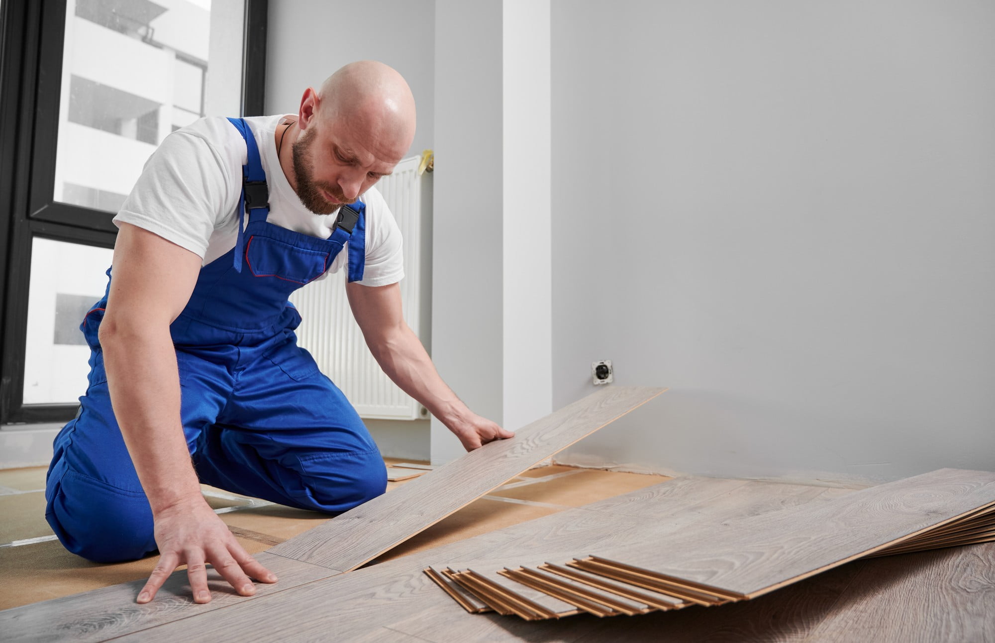 Sure! Here’s a detailed alt text description for the image:"A man dressed in dark blue workwear, including a matching jacket and trousers, is installing laminate flooring in a well-lit room. He is kneeling on the floor, carefully positioning a piece of laminate plank into place. His tool belt, which has several compartments and tools, is visible around his waist. In the background, the room appears to be freshly painted with light-coloured walls, and there is a large window that lets in natural light, illuminating the work area. The man’s focused expression suggests he is diligently paying attention to the alignment and fit of the laminate pieces. The floor around him is scattered with various tools and additional planks of laminate, indicating an ongoing flooring project."