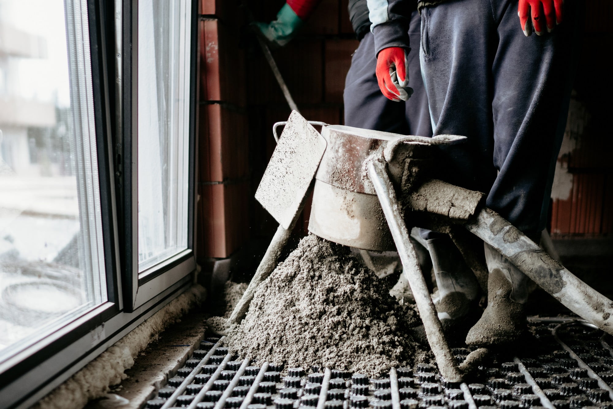 The image depicts a close-up view of a construction scene where a worker is leveling a freshly laid sand and cement screed floor. The screed mixture appears smooth and damp, indicating that it has been recently spread. The worker’s hands, clad in construction gloves, are holding a long, straight-edged trowel or screed board, which they are using to level the surface meticulously. The background shows a partially constructed building with unfinished walls, and the floor space is expansive, suggesting a large area being worked on. The lighting is natural, likely from openings or windows not visible in the image. Various construction tools and materials can be seen scattered around, giving the setting an authentic, in-progress feel. The overall focus is on the precise and skilled process of ensuring the screed is level and smooth, essential for a quality finish in flooring.