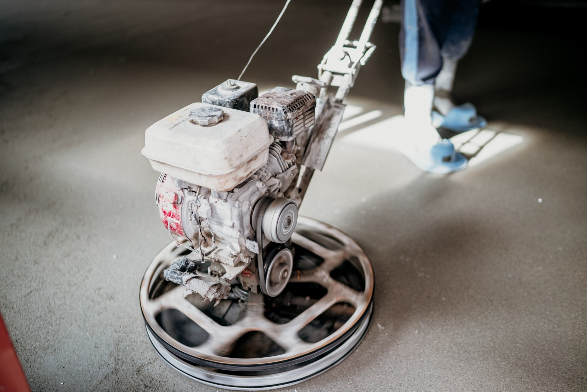 The image features a construction worker operating a power trowel to smooth and finish a large concrete surface. The worker is wearing a safety helmet, a bright high-visibility vest, long pants, and sturdy work boots. They are standing on the freshly poured concrete while maneuvering the power trowel, which has four wide, spinning blades underneath a protective guard. The surrounding environment appears to be a construction site with an overcast sky. In the background, there are various construction materials and equipment, including wooden planks, orange safety barriers, and steel rebar. The image emphasizes the precision and skill required in the concrete finishing process.