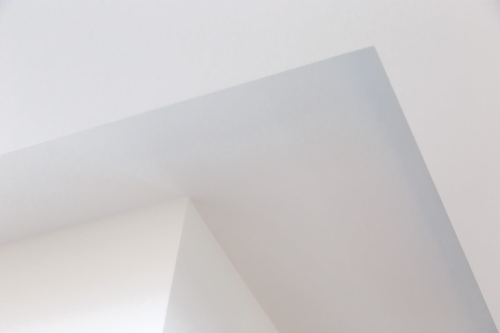 The image shows a bottom-view perspective of a wall corner, focusing on its intersection with a ceiling. Both the wall and the ceiling are constructed from smooth, white plasterboard, creating clean, sharp lines where they meet. The plasterboard appears to be freshly installed or well-maintained, with no visible damage, stains, or irregularities. The image is likely taken during the construction or interior finishing phase of a building project, emphasising the pristine condition and accuracy of the plasterboard work. The lighting in the photo highlights the even texture and seamless joins, contributing to a minimalist and modern aesthetic.