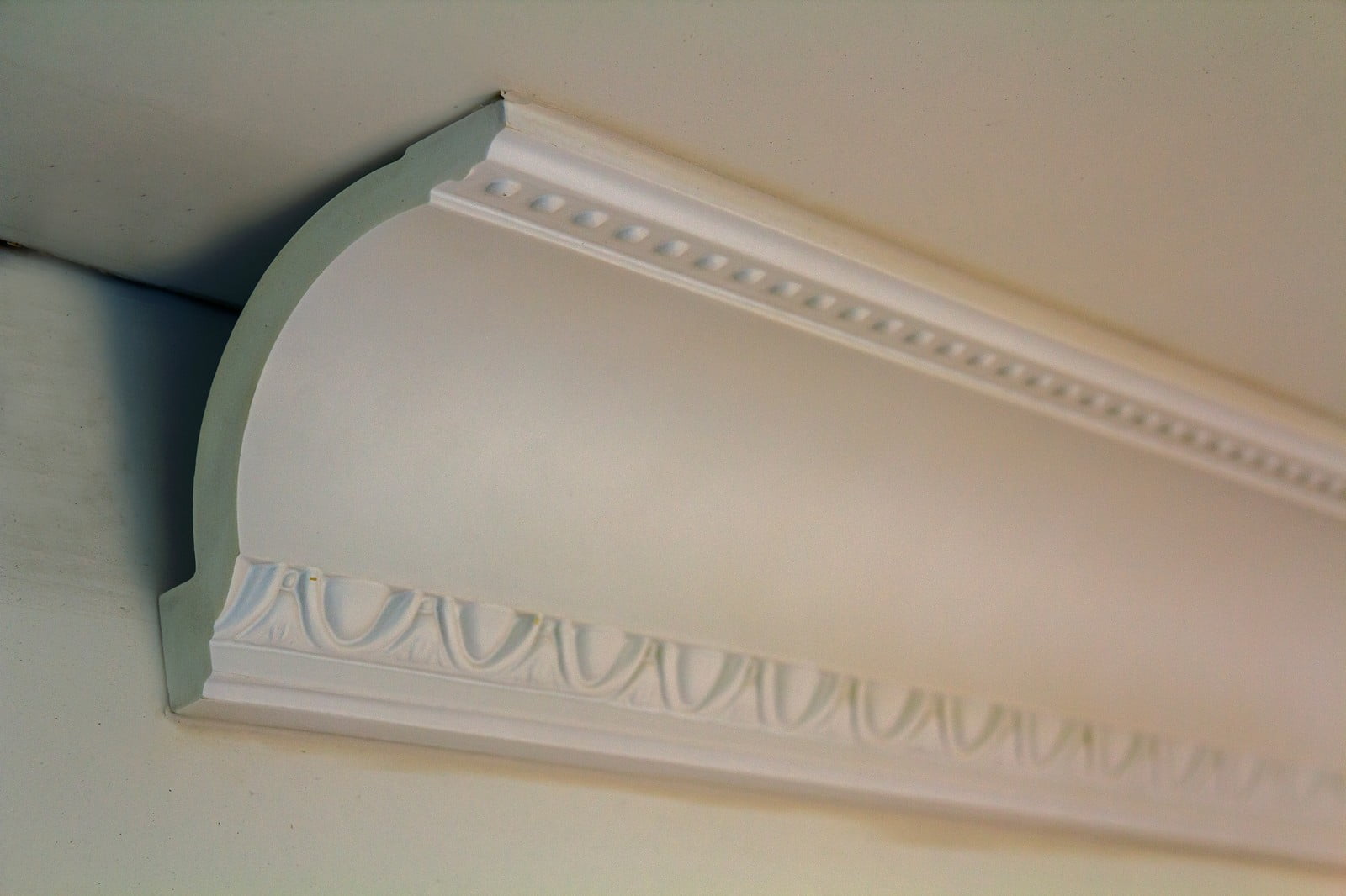 The image is a close-up photograph showcasing a detailed section of white molding in an interior setting. The molding displays an intricate design featuring classical, ornamental patterns typical of traditional architectural decoration. The crisp white colour of the molding contrasts elegantly against the slightly darker background, drawing attention to the fine craftsmanship. The lighting in the image highlights the depth and texture of the molding’s design, emphasising the relief work and shadows created by its intricate patterns. The overall aesthetic conveys a sense of sophistication and attention to detail, reflecting high-quality interior decor.