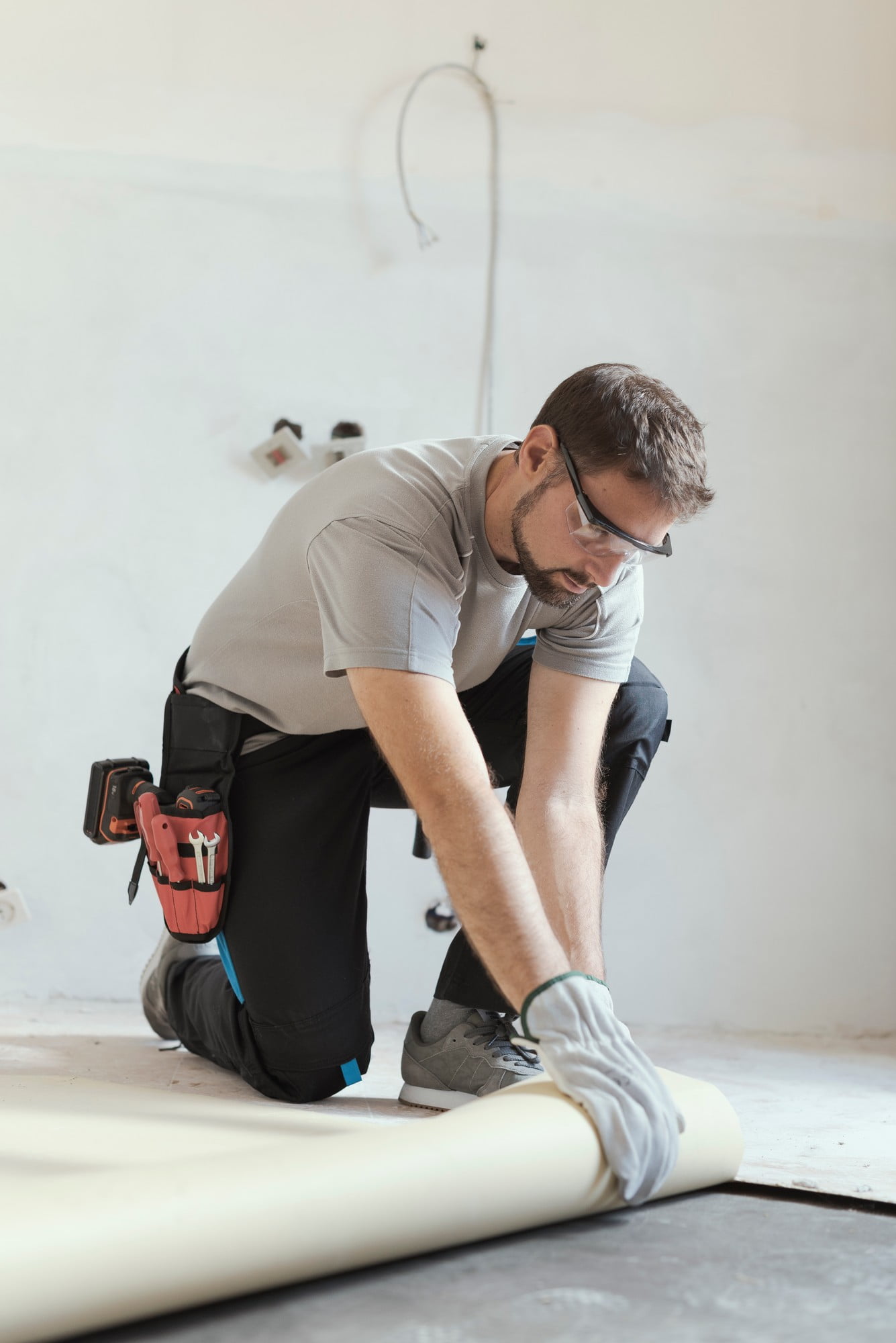 **Alt Text: Contractor Removing Old Linoleum Flooring**In the image, a contractor is engaged in the process of removing old linoleum flooring. The scene is set in a room with bare, unfinished walls and a partially removed linoleum floor, revealing the subfloor beneath. The contractor is wearing sturdy work attire, including a pair of heavy-duty gloves and protective knee pads. He is crouched down on one knee, firmly gripping the edge of the linoleum with one hand while the other hand holds a large floor scraper tool, used to pry up the linoleum. The ambient light in the room is natural, suggesting daytime work, and the floor appears to be in the early stages of removal, with chunks of linoleum already peeled away and scattered around. In the background, various tools and supplies are visible, hinting at the ongoing renovation work. The room has an industrial look, with exposed elements and a focus on practical functionality over finished aesthetics. The image captures the manual labour and effort involved in floor renovation.