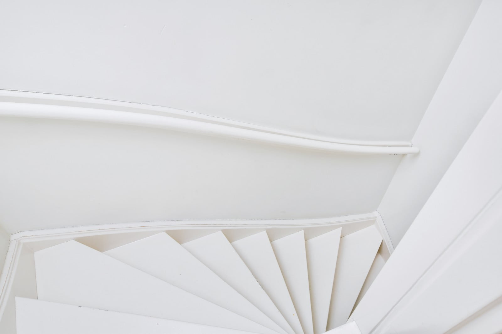 The image depicts a highly modern and stylish white spiral staircase situated in a contemporary flat. The architectural design of the staircase is sleek and minimalist, featuring smooth, flowing lines that create a seamless and elegant spiral. The white colour of the staircase enhances the sense of space and light within the room, contributing to an airy and open ambiance. The staircase is constructed with a central metal axis around which the stairs twist gracefully, leading up to another level in the flat.To the left of the staircase, there is a glass railing that encases the upper floor, offering safety without detracting from the open-plan layout. The floor is finished with light-coloured wood, complementing the modern aesthetic of the space. Large, floor-to-ceiling windows flank the room, flooding it with natural light and providing expansive views of the urban landscape outside. The walls are painted in a crisp white, maintaining the clean aesthetic and making the entire area feel cohesive and expansive.Furnishings in the flat are minimal yet stylish, consistent with the overall contemporary décor. A sleek, low-profile sofa in a neutral tone is positioned in the bottom right corner of the image, paired with a modern rectangular coffee table and a plush area rug. The overall environment exudes a sense of luxury and sophistication through its combination of modern architectural elements, strategic use of lighting, and minimalist yet elegant decor.